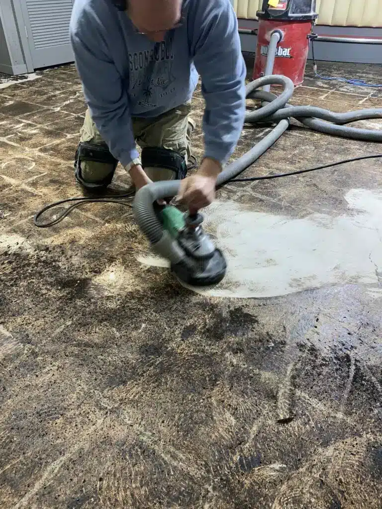 A person is kneeling on a concrete floor, using a grinder attached to an industrial vacuum, possibly in the process of floor refinishing or preparation.