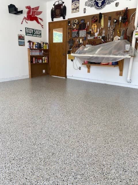 A clean, organized garage with speckled flooring, various wall-mounted tools, signage, and a wood door. A classic car is partially visible under a cover.