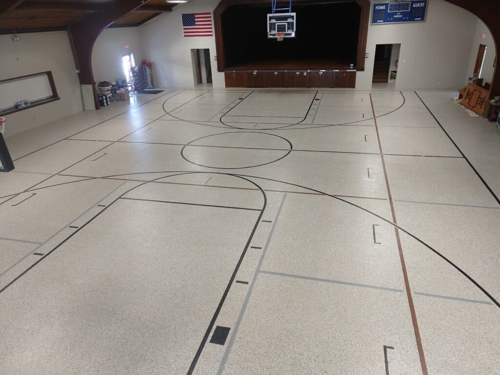 An indoor basketball court with lines marked for play, featuring a basketball hoop and a scoreboard. An American flag hangs on the wall.
