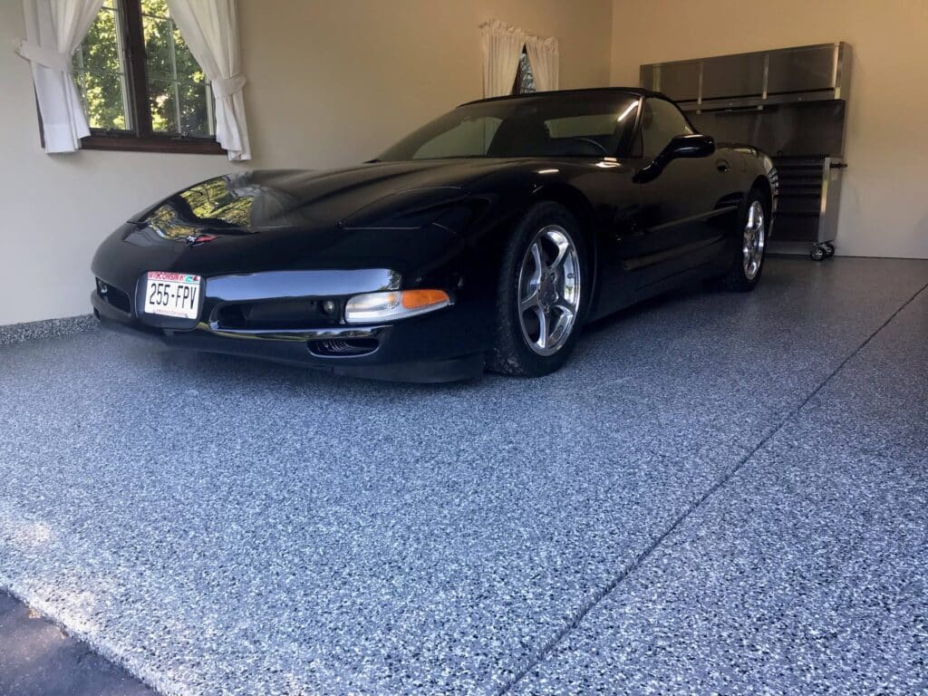 A sleek black sports car parked in a garage with a speckled blue floor, near a stainless steel tool chest, in a clean and organized space.