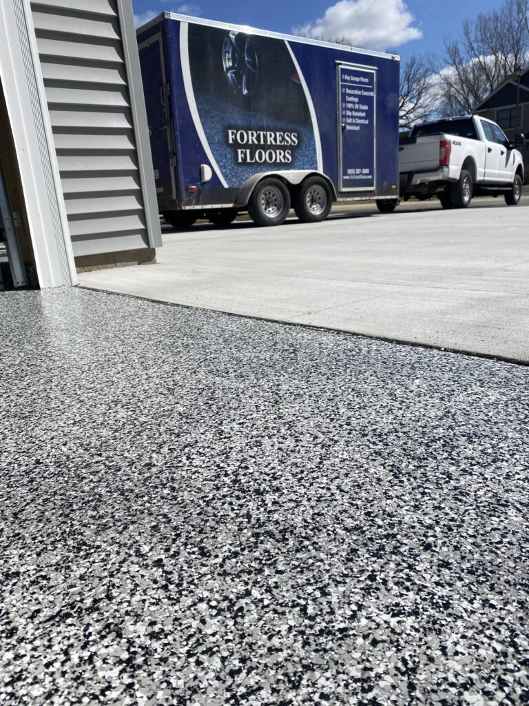 The image shows a close-up of a textured floor leading to a parked trailer labeled "FORTRESS FLOORS" with a pickup truck in the background under a clear sky.