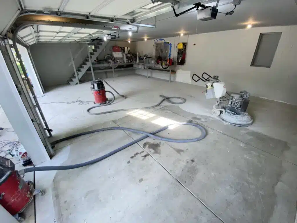 A spacious, unoccupied garage with an open door, a staircase, various equipment, and ongoing floor resurfacing work using a concrete grinder and vacuum.