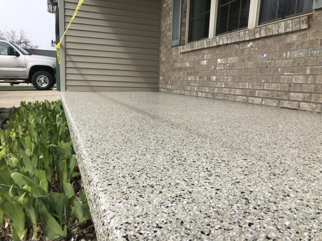 A textured concrete walkway leads to a house with brick siding; green plants line one side, and a parked truck is visible in the distance.