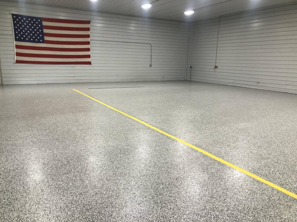 A spacious garage with a speckled epoxy floor and a large American flag hung on the back wall above a yellow line. The space appears clean and well-maintained.