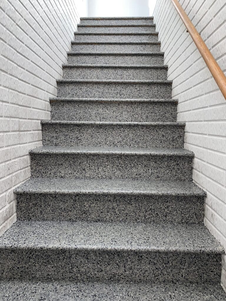 A straight flight of granite stairs with a wooden handrail on the right and white brick walls on both sides, leading upwards.