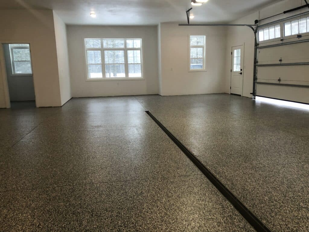 Empty garage with speckled epoxy flooring, white walls, multiple windows for natural light, a closed overhead door, and an entry door.