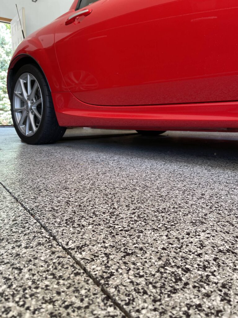 A close-up side view of a red vehicle parked on a textured gray surface. The focus is on the car's rear side panel and wheel.